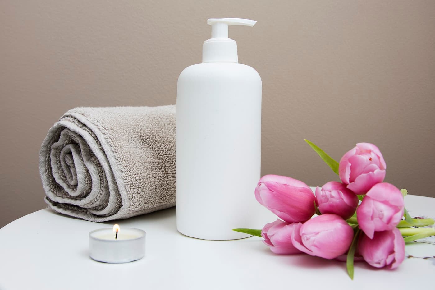 A rolled towel, a lotion dispenser, a lit candle and tulips on a spa table.